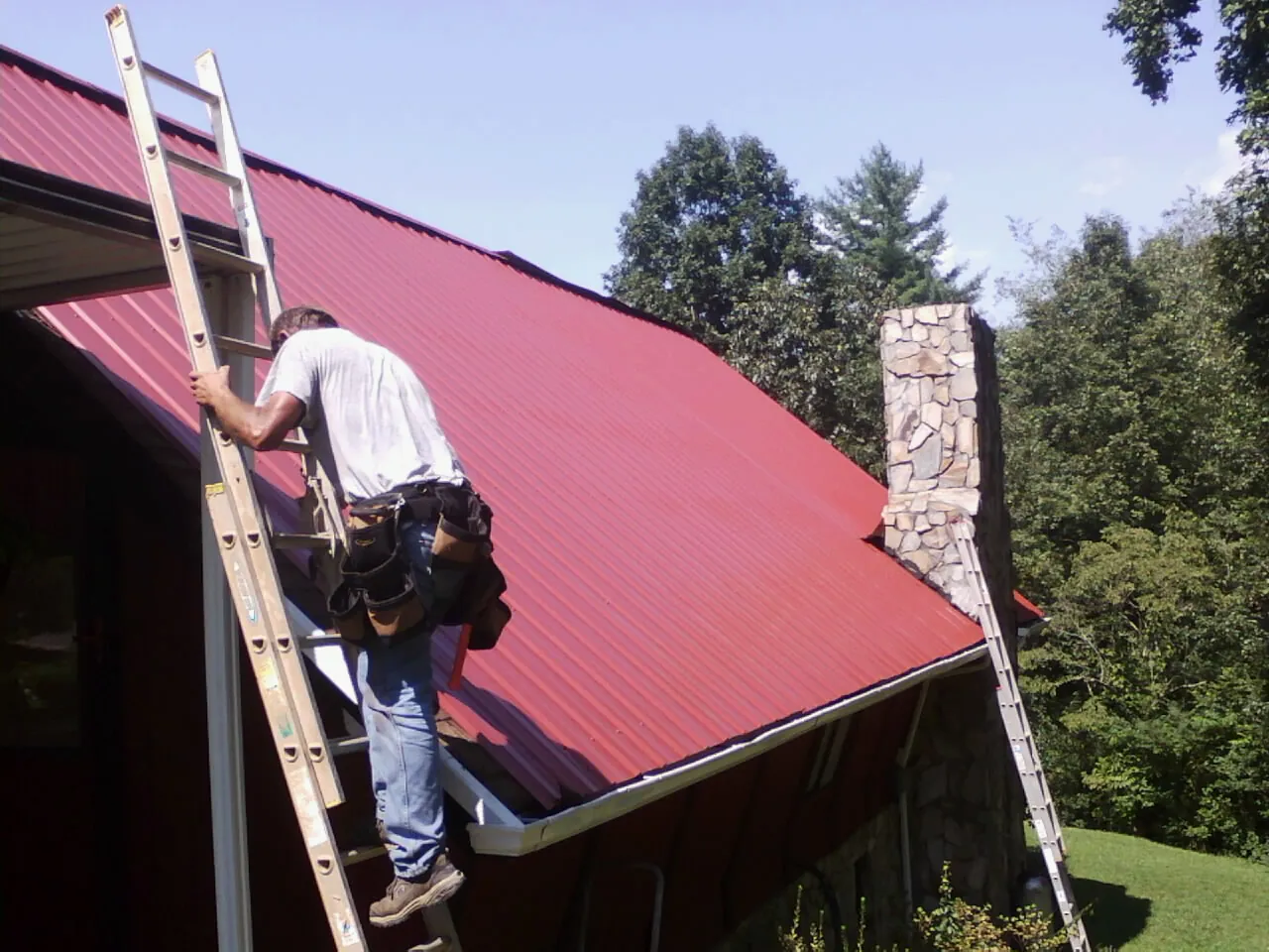 Roofer on a ladder beside a red metal roof and stone chimney — Soffit & Fascia Repair by Roofing Brothers in Napanoch